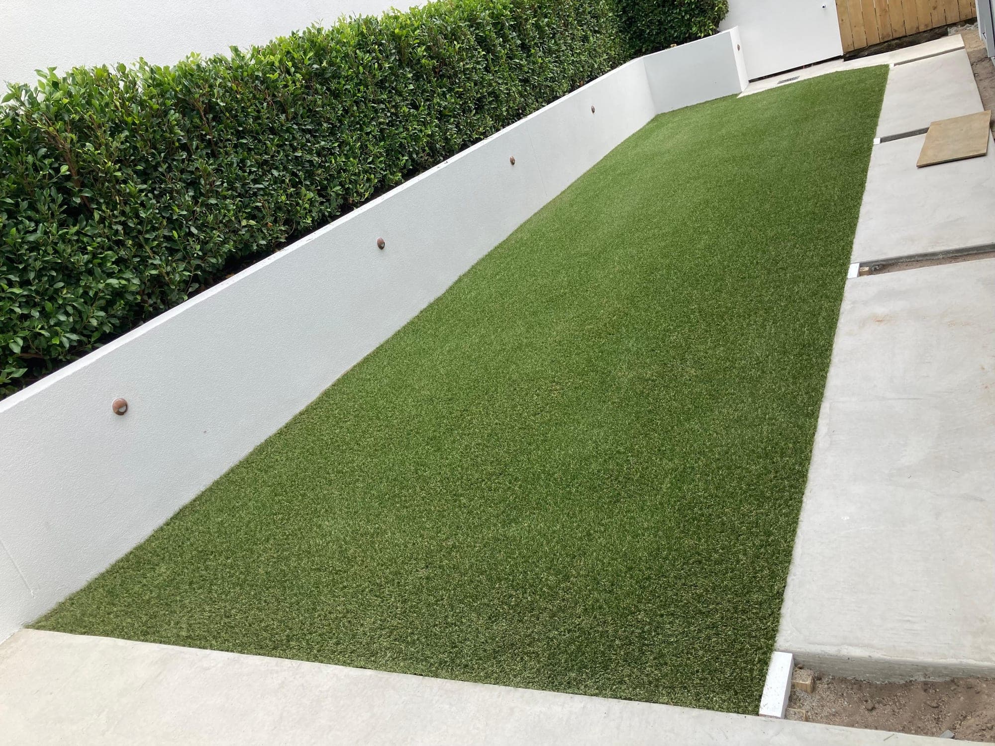 Artificial turf installed in a long strip between a white plastered wall and concrete paving