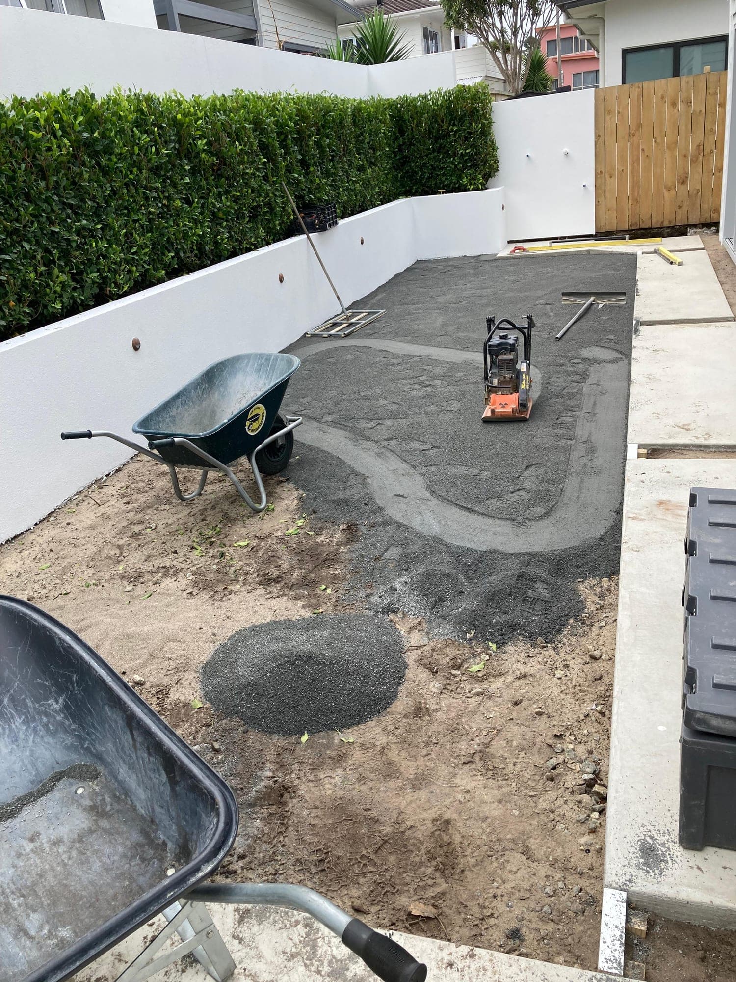 Plate compactor working a basecourse in a narrow courtyard beside a white plastered wall and clipped hedge