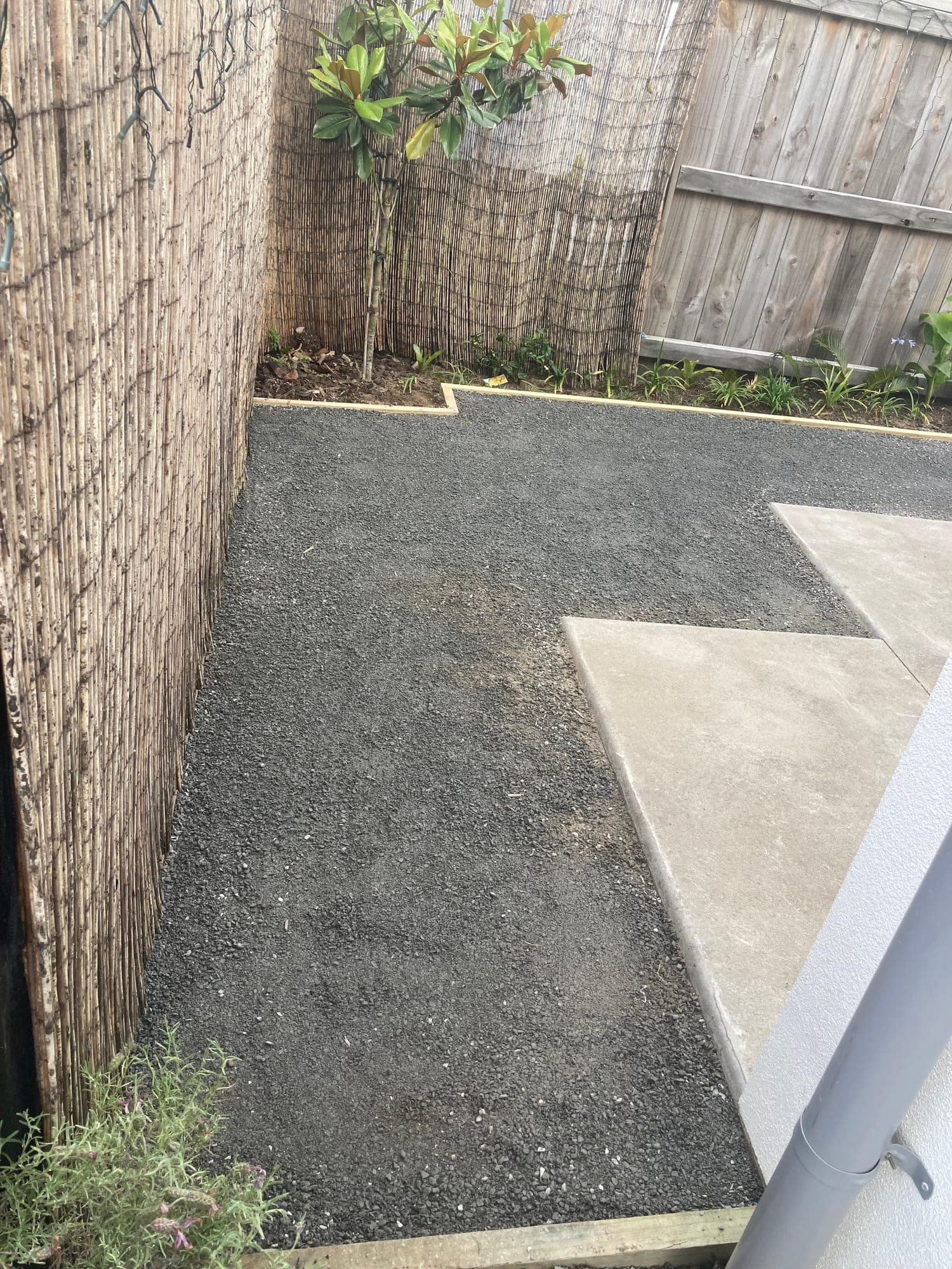 Compacted basecourse in a narrow garden run beside a reed screen fence and concrete pavers