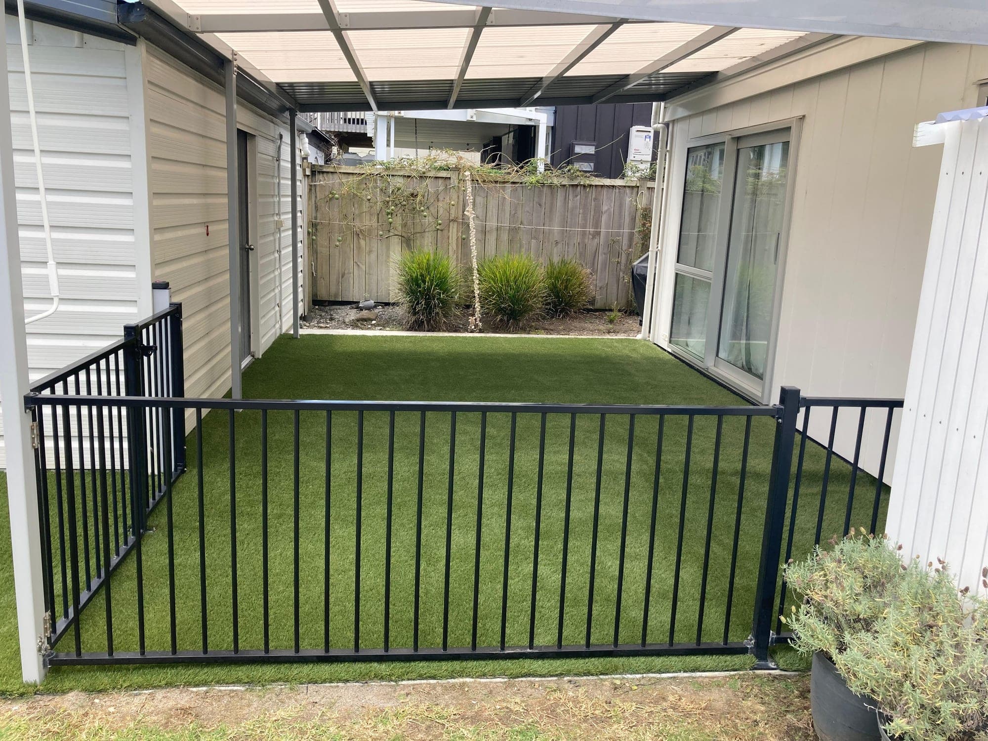 Artificial turf inside a black aluminium pool fence, running under a pergola beside a house